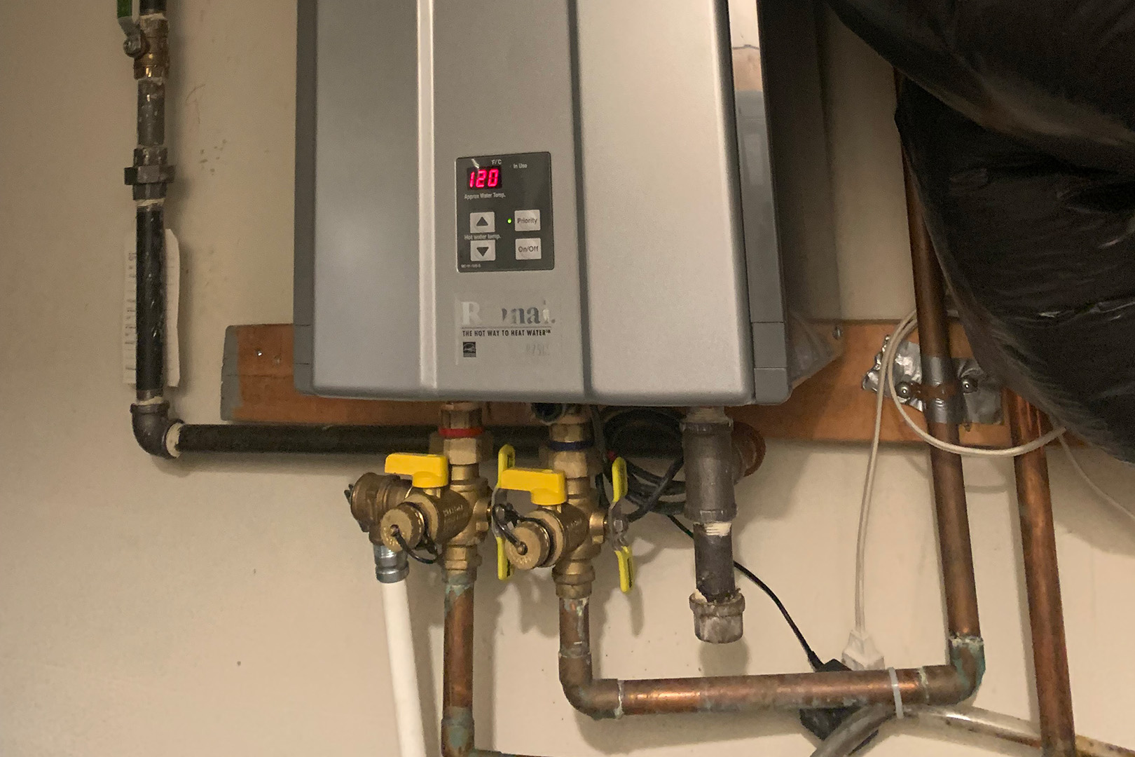 Installed Rinnai tankless water heater with digital temperature display mounted on wall, showing copper pipe connections and yellow shut-off valves beneath unit