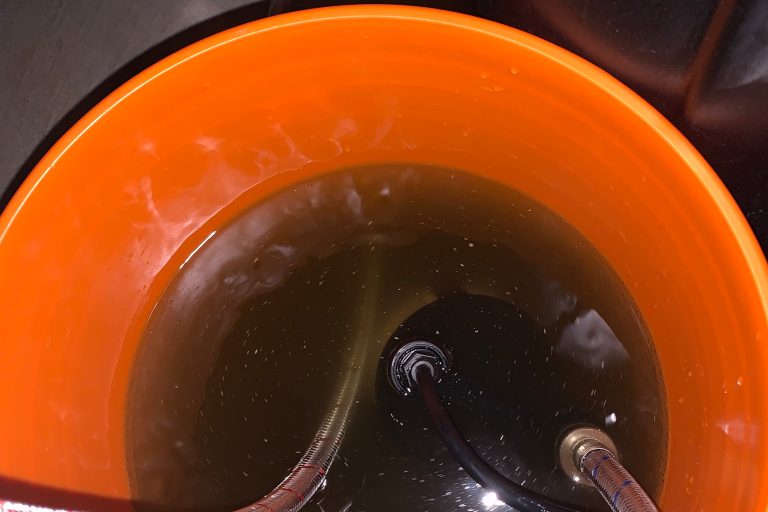 View inside an orange bucket showing dark, sediment-filled water during a tankless water heater flush maintenance procedure